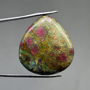 May include: A teardrop-shaped gemstone with a mix of colors. The stone has a base of green and gold with patches of deep red. The stone is held by a pair of silver tweezers, showcasing its polished surface and unique color variations.