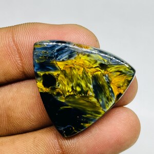 May include: A polished, triangular gemstone with a striking pattern of swirling blue, yellow, and black colors. The stone's surface is smooth and reflective, showcasing the natural variations in color and texture. The gemstone is held in a hand.