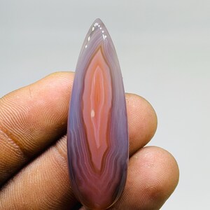 Puede incluir: Una gema pulida en forma de lágrima con un patrón de colores en capas. La piedra presenta bandas de púrpura, naranja y blanco, creando un diseño único y natural. La gema se sostiene en una mano.