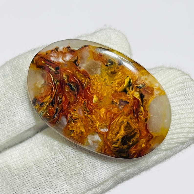 May include: An oval-shaped gemstone with a translucent base, showcasing a vibrant display of golden, orange, and red hues. The stone's internal patterns create a dynamic, swirling effect, with areas of deep color and lighter, more transparent sections. The polished surface reflects light.