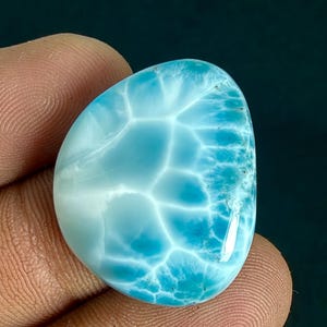 May include: A polished, teardrop-shaped Larimar gemstone featuring a vibrant sky-blue color with intricate, web-like white patterns across its smooth surface. The stone displays a high-gloss finish that reflects light, highlighting the natural variations in the blue and white mineral structure throughout the piece.