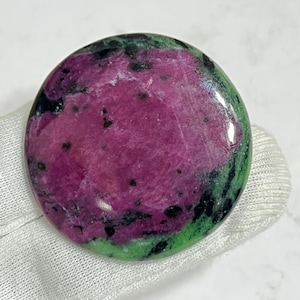 May include: A polished, round gemstone with a vibrant color palette. The center features a deep magenta hue, speckled with black inclusions. A band of green surrounds the magenta, with a dark green outer edge. The stone is set on a white surface.