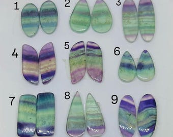 May include: A collection of nine pairs of polished fluorite gemstones in various shapes. The stones display a range of colors, including purple, green, and blue, with banded patterns. Each pair is numbered for identification.