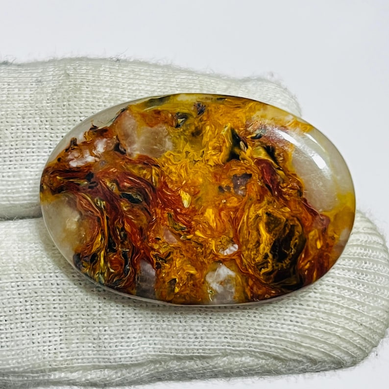 May include: Oval-shaped gemstone with a swirling pattern of gold, orange, and brown hues. The polished stone has a translucent appearance with a mix of colors and textures, creating a unique and natural design. The gemstone is set against a white background.