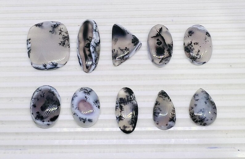 May include: A collection of polished dendritic agate gemstones in various shapes, including oval, teardrop, and triangular. The stones feature a translucent, milky white base with intricate black and gray dendritic patterns.