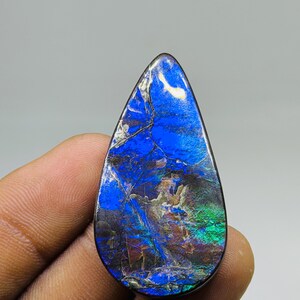 Può includere: Una gemma a forma di goccia con una vibrante esposizione di colori. La pietra presenta una base blu scuro con striature verdi, marroni e argentate, creando un motivo iridescente unico. La superficie lucida riflette la luce.