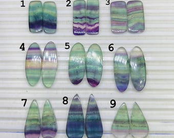 May include: A collection of nine pairs of polished fluorite gemstones in various shapes. The stones display a range of colors, including green, purple, and blue, with visible banding patterns. The shapes include rectangles, ovals, and teardrops.