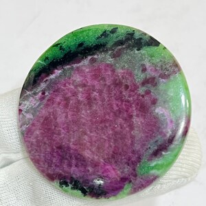 May include: A polished, oval-shaped gemstone featuring a vibrant mix of colors. The stone showcases a deep magenta center, encircled by a band of bright green and speckled with black inclusions. The surface is smooth and reflective.