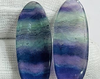 Puede incluir: Dos piedras preciosas de fluorita de forma ovalada. Las piedras muestran un patrón en capas de tonos azules, morados y verdes. Las superficies pulidas son lisas y reflectantes, mostrando las variaciones naturales de color.
