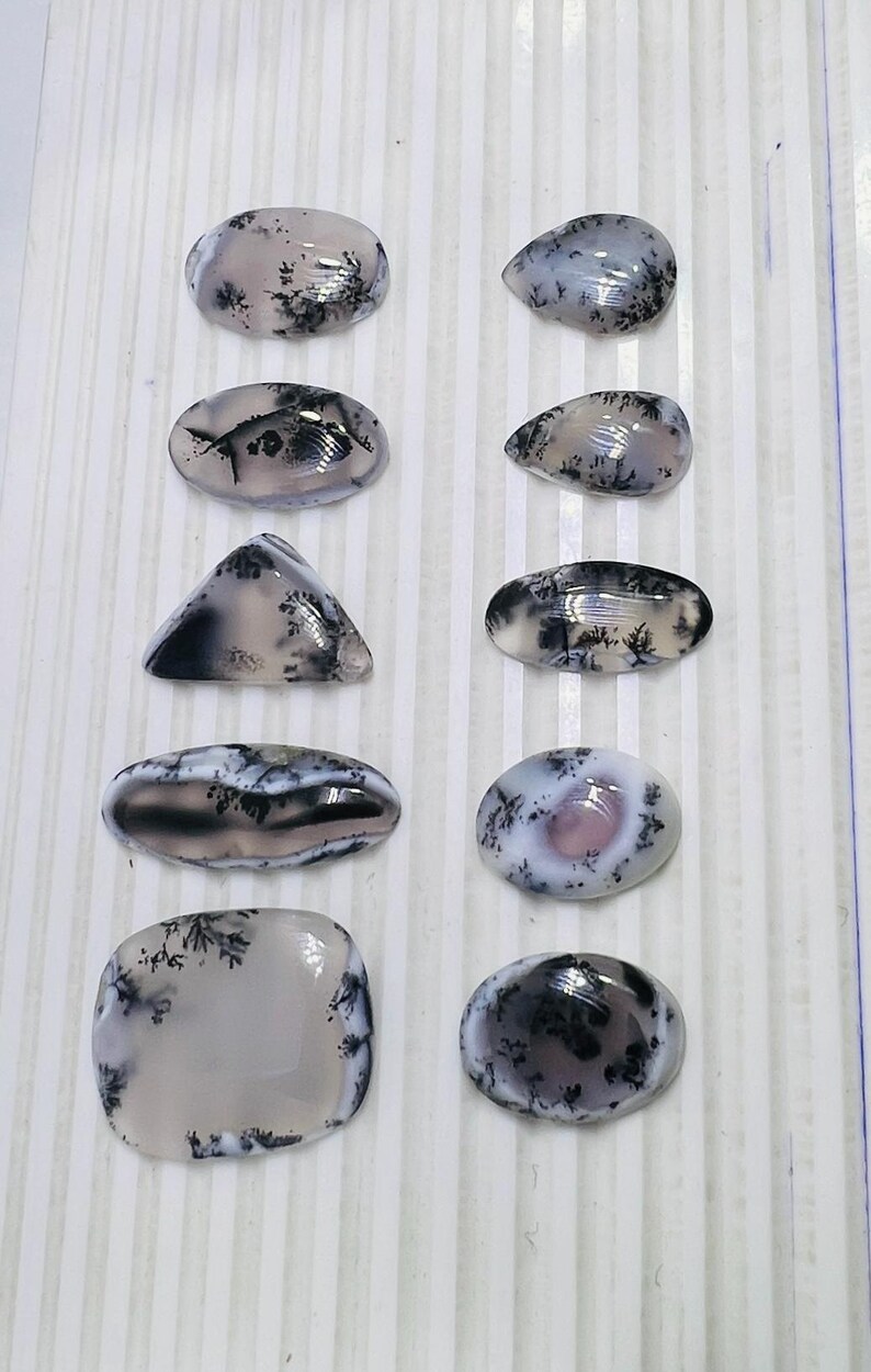 May include: A collection of polished dendritic agate gemstones in various shapes, including oval, teardrop, and triangular. The stones feature a translucent base with black and gray fern-like inclusions. The gemstones are displayed on a white striped surface.