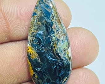 May include: A teardrop-shaped gemstone with a polished surface. The stone features a swirling pattern of deep blue, yellow, and brown hues. The gemstone is held between fingers, showcasing its size and shape.