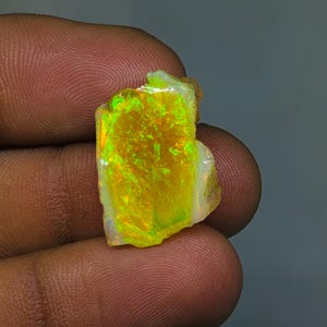 Puede incluir: Una vibrante piedra preciosa de ópalo en bruto, que muestra un espectro de colores. La piedra muestra una base amarilla brillante con destellos de verde y naranja, sostenida entre los dedos. La superficie del ópalo tiene un aspecto natural y texturizado.