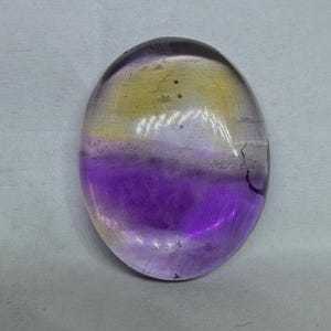 Puede incluir: Una gema ovalada con un degradado de colores. La parte superior es amarilla, pasando a una banda blanca y luego a un púrpura vibrante en la parte inferior. La piedra tiene un aspecto pulido y translúcido.