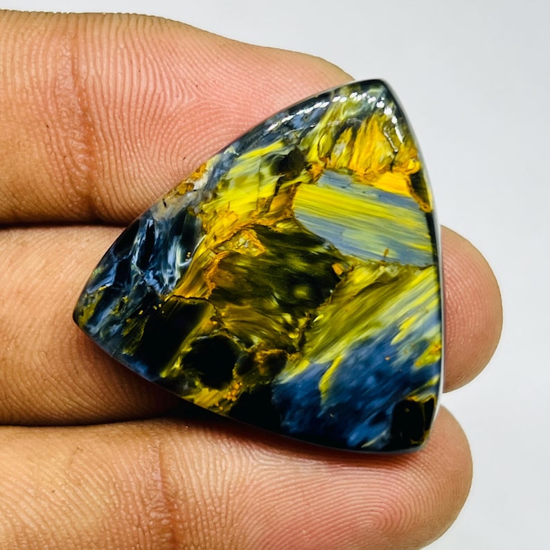 May include: A triangular gemstone with a polished surface. The stone features a striking blend of colors, including deep blues, vibrant yellows, and hints of black. The gemstone's unique pattern and color variations create a visually appealing aesthetic.