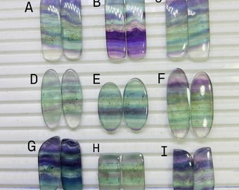 May include: A collection of nine pairs of polished gemstones in various shapes and colors. The stones display a range of hues, including purple, green, and clear, with visible banding patterns. The gemstones are arranged in rows for display.
