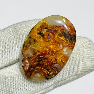 May include: Oval-shaped gemstone with a polished surface. The stone features a swirling pattern of orange, yellow, brown, and black hues. The gemstone is set against a white background.
