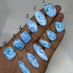 May include: A collection of twelve polished larimar gemstones in various shapes, including oval, rectangular, and teardrop. The stones display a light blue color with white marbling patterns, held in a hand against a neutral background.