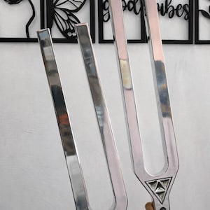 May include: Two silver tuning forks are held up against a white wall with black framed wall art. The tuning forks have a triangular base and a long, rectangular shape. The wall art includes a butterfly and the words "good vibes".
