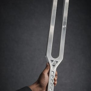 Może przedstawiać: Duży, srebrny kamerton na ciemnoszarym tle. Kamerton ma tekst "174 HZ Giant Fork" w lewym górnym rogu. Podstawa kamertonu ma szereg małych, okrągłych otworów.