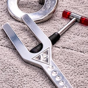 Può includere: Un set di utensili in metallo argentato, tra cui un pezzo circolare con un design decorativo, uno strumento a forcella con un emblema triangolare e un martello con manico rosso. Gli utensili sono disposti su una superficie strutturata grigio chiaro.