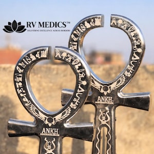 May include: Two silver-colored Ankh pendants with engraved hieroglyphs and decorative patterns. The pendants have an oval top and a cross-like base. The text "ANKH" and "RV MEDICS" are visible.