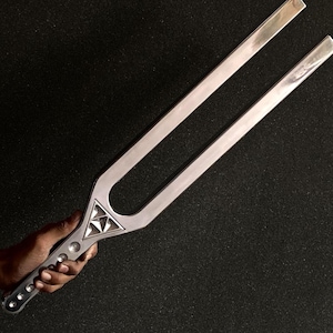May include: A silver tuning fork with a decorative triangular design, held against a dark background. The tuning fork has a handle with several small, round indentations. The tines are parallel and smooth.