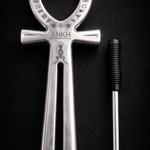 May include: A silver-colored Ankh symbol with etched details and the word "ANKH" engraved. A black-handled tool with a silver shaft is positioned next to the Ankh. The background is a dark, textured surface.