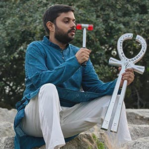 May include: A person holding a silver ankh and a red and silver hammer. The ankh is a large, ornate symbol with hieroglyphic-like markings. The hammer has a red head and a silver handle. The person is wearing a blue tunic and white pants.