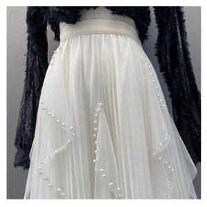 May include: A white tulle skirt with pearl and rhinestone accents. The skirt has a layered design and is paired with a black, fringed top. The skirt's waistline is accented with a cream-colored band.