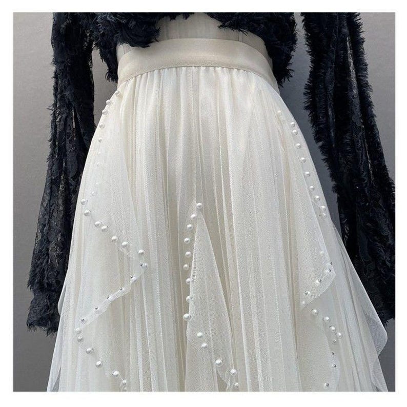 May include: A white tulle skirt with pearl and rhinestone accents. The skirt has a layered design and is paired with a black, fringed top. The skirt's waistline is accented with a cream-colored band.