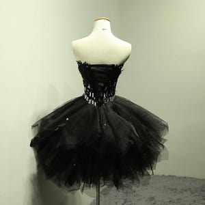May include: A black strapless dress on a mannequin. The bodice features a corset design with feather accents and a sparkling belt. The skirt is a layered tulle, creating a voluminous, short silhouette. The dress is displayed on a metal stand.