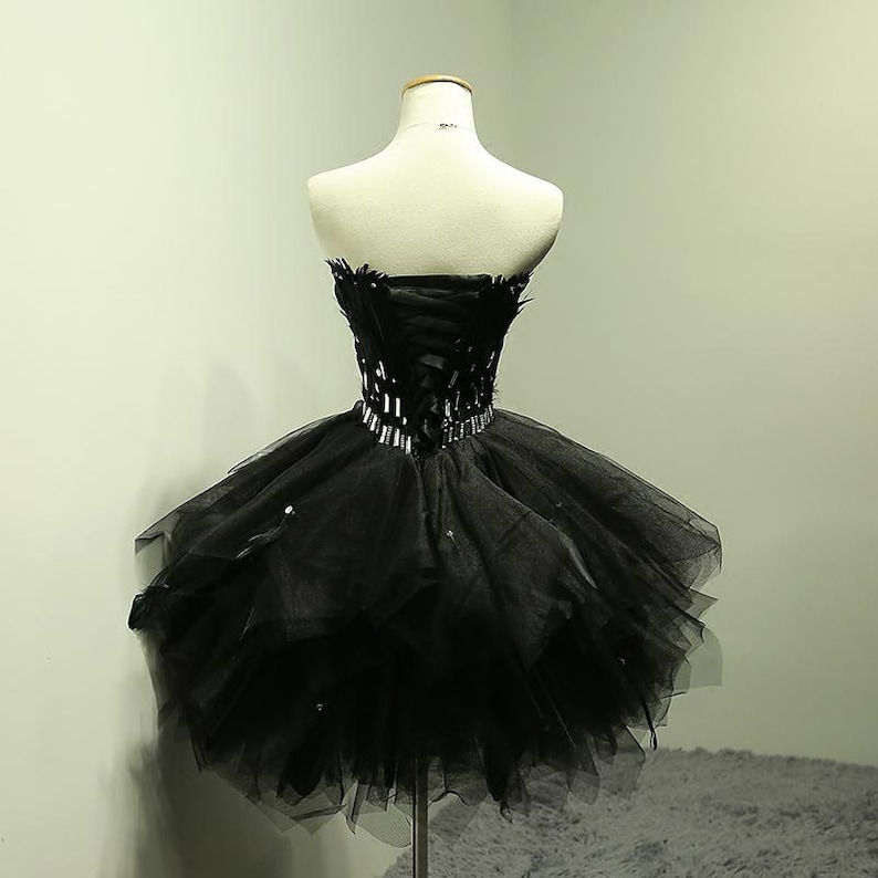 May include: A black strapless dress on a mannequin. The bodice features a corset design with feather accents and a sparkling belt. The skirt is a layered tulle, creating a voluminous, short silhouette. The dress is displayed on a metal stand.