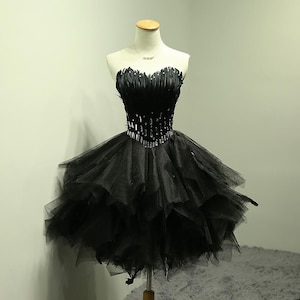 May include: A strapless black cocktail dress on a mannequin. The bodice is embellished with feathers and rhinestones, and the skirt is layered tulle. The dress is designed to be knee-length, offering a classic and elegant look.