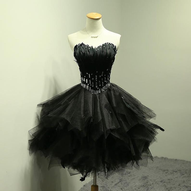 May include: A strapless black cocktail dress on a mannequin. The bodice is embellished with feathers and rhinestones, and the skirt is layered tulle. The dress is designed to be knee-length, offering a classic and elegant look.