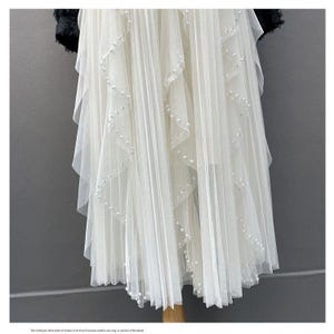 May include: A white, layered tulle skirt with delicate pearl accents. The skirt features a pleated design and a flowing silhouette. The top portion of the skirt is partially obscured by a black, textured fabric.