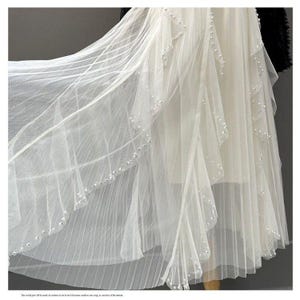 May include: A flowing white dress with layers of sheer fabric. The dress features delicate pearl embellishments along the edges of the layers, creating a subtle shimmer. The design suggests a formal or bridal theme.