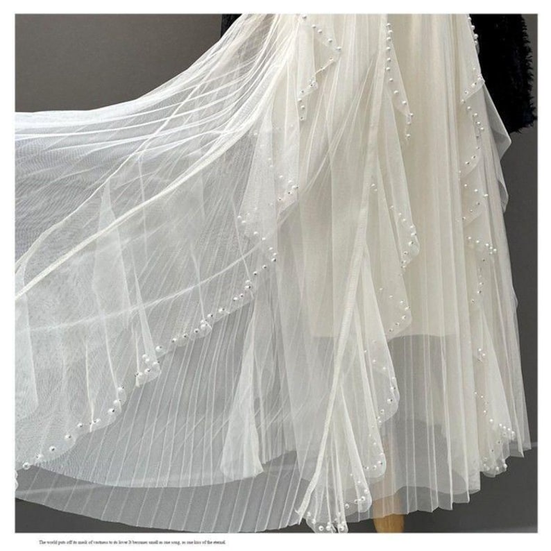 May include: A flowing white dress with layers of sheer fabric. The dress features delicate pearl embellishments along the edges of the layers, creating a subtle shimmer. The design suggests a formal or bridal theme.
