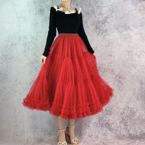 Op de afbeelding: Een zwarte top met lange mouwen en witte details, gecombineerd met een rode tule rok. De rok is gelaagd en wijd uitlopend, met een gefranjerde zoom. De outfit wordt gecompleteerd met sandalen met hakken.