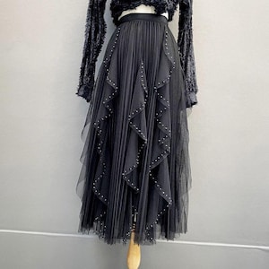 May include: A black tulle skirt with pleated detailing and layers of cascading ruffles. The skirt is adorned with small, silver-colored studs. The skirt is part of a two-piece outfit, with a black, fuzzy long-sleeved top.