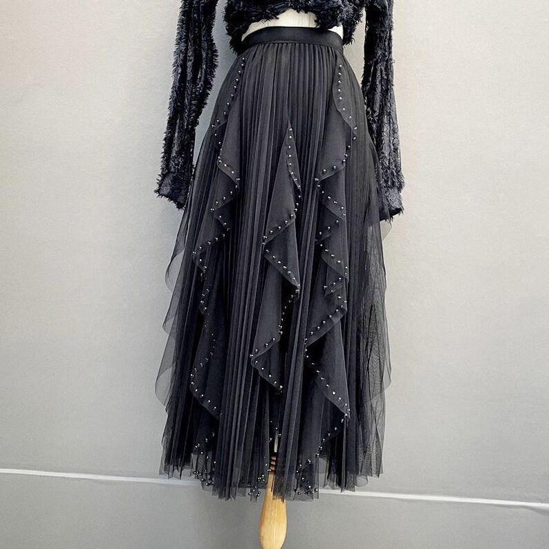 May include: A black tulle skirt with pleated detailing and layers of cascading ruffles. The skirt is adorned with small, silver-colored studs. The skirt is part of a two-piece outfit, with a black, fuzzy long-sleeved top.