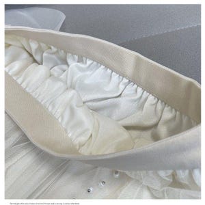 May include: Close-up of a white tulle skirt with an elastic waistband. The skirt features multiple layers of soft, flowing fabric and is adorned with small, shimmering embellishments. The waistband is a neutral beige color.