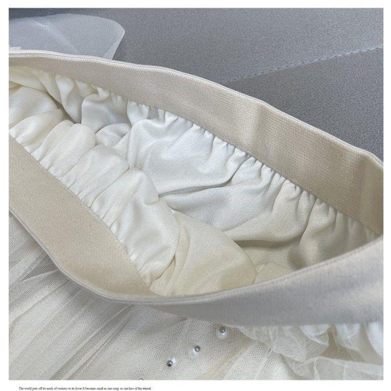 May include: Close-up of a white tulle skirt with an elastic waistband. The skirt features multiple layers of soft, flowing fabric and is adorned with small, shimmering embellishments. The waistband is a neutral beige color.