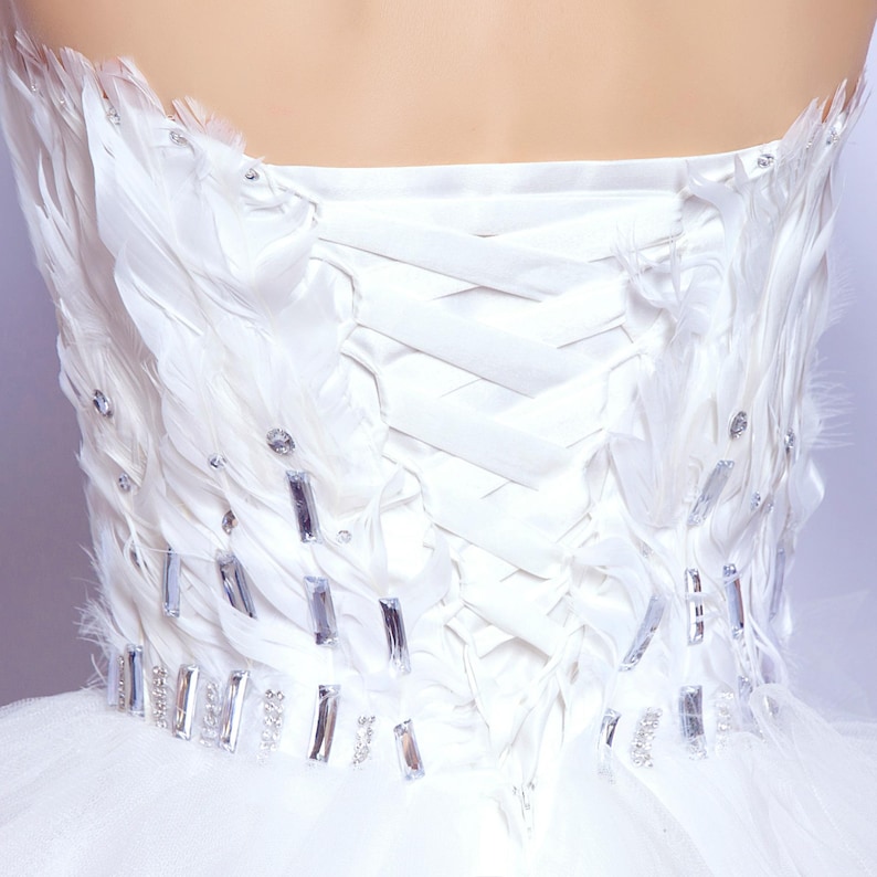 May include: A white strapless dress bodice with feather and crystal embellishments. The bodice features a corset back with satin ribbons and is adorned with white feathers and rectangular crystals. The dress appears to be made of tulle.