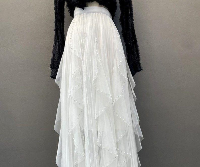 May include: A white tulle skirt with a layered design and beaded accents. The skirt is paired with a black, fuzzy top. The skirt has a flowing, elegant appearance, suitable for special occasions.