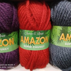 May include: Three balls of James C. Brett Amazon Super Chunky yarn in shades of purple, red, and grey. Each ball has a green label with the brand name and product details. The yarn is thick and appears soft.