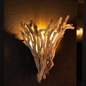 Puede incluir: Un aplique de pared construido con un conjunto de ramas de madera flotante clara y desgastada. La disposici&oacute;n de la madera crea una forma de abanico, con una luz c&aacute;lida y dorada que emana desde detr&aacute;s y entre las ramas.