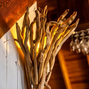 May include: A rustic wall sconce crafted from natural driftwood. The light fixture features numerous pieces of weathered wood arranged to create a unique, organic design. The warm light shines through the gaps in the wood, creating a cozy ambiance.