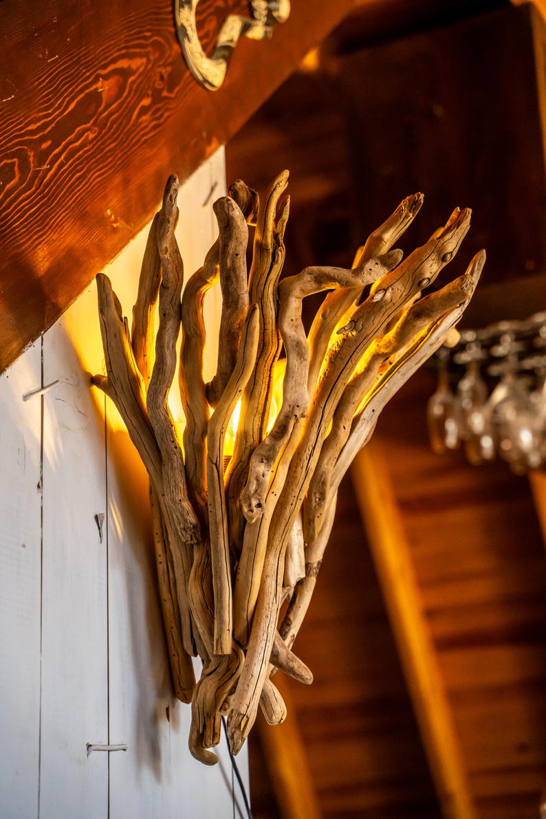 May include: A rustic wall sconce crafted from natural driftwood. The light fixture features numerous pieces of weathered wood arranged to create a unique, organic design. The warm light shines through the gaps in the wood, creating a cozy ambiance.