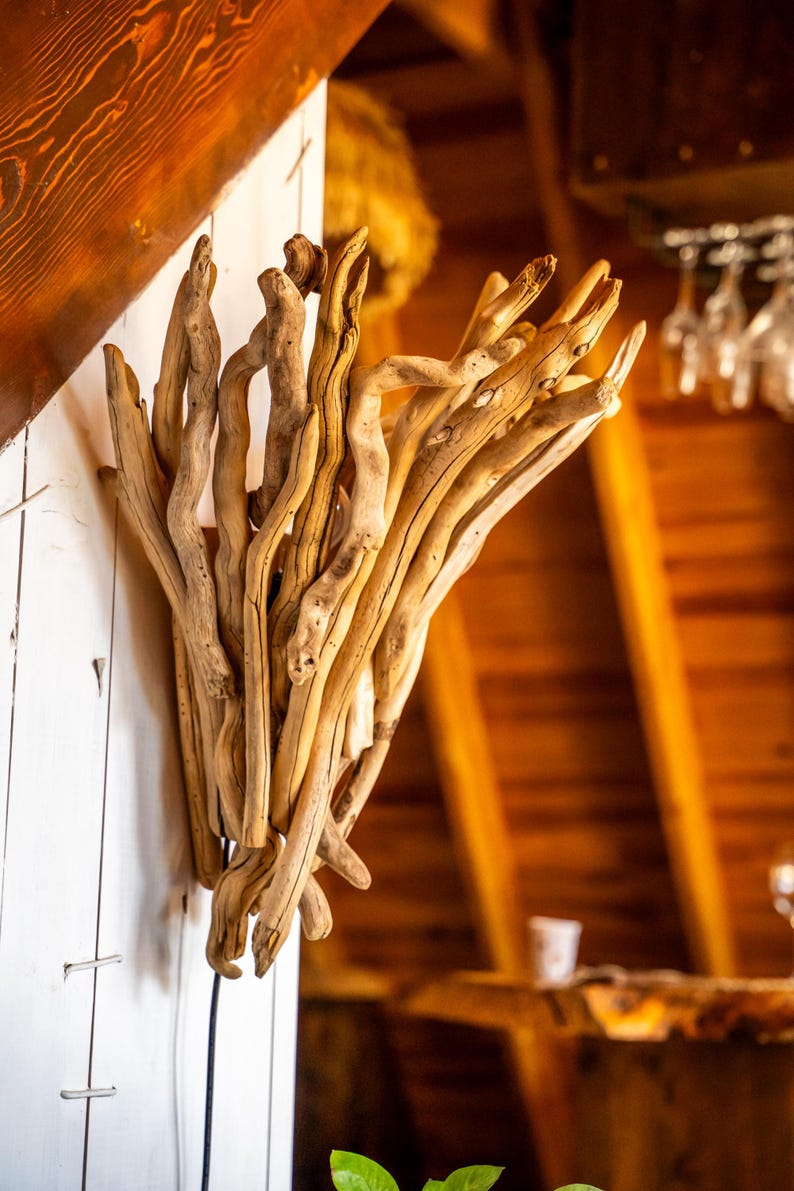 Puede incluir: Un aplique de pared construido con un grupo de ramas de madera flotante de color marr&oacute;n claro y desgastadas, dispuestas en una formaci&oacute;n vertical en forma de abanico. Las piezas de madera natural y nudosa est&aacute;n unidas en una estructura org&aacute;nica.