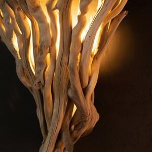 Puede incluir: Un aplique de pared construido con un conjunto de ramas de madera flotante de color claro y aspecto desgastado. La disposici&oacute;n de la madera crea una forma vertical en abanico, con una luz c&aacute;lida y dorada que emana entre las ramas.